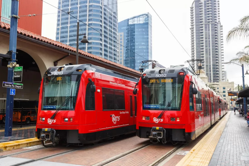 Two red San Diego trolley cars at Santa Fe Depot station with high-rise buildings in background.